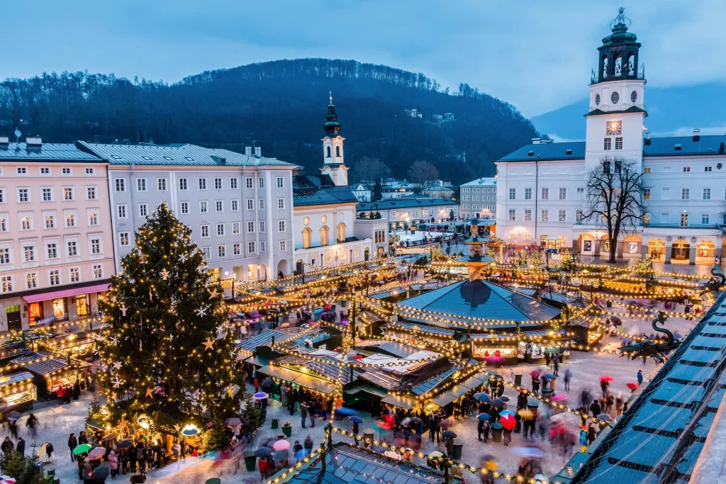 The image shows the Salzburg Christmas market. The center of the image shows the booths of the artisans selling their wares surrounded by a church of the city square. The image is taken at sunset, providing a profile of the surrounding high ground.    